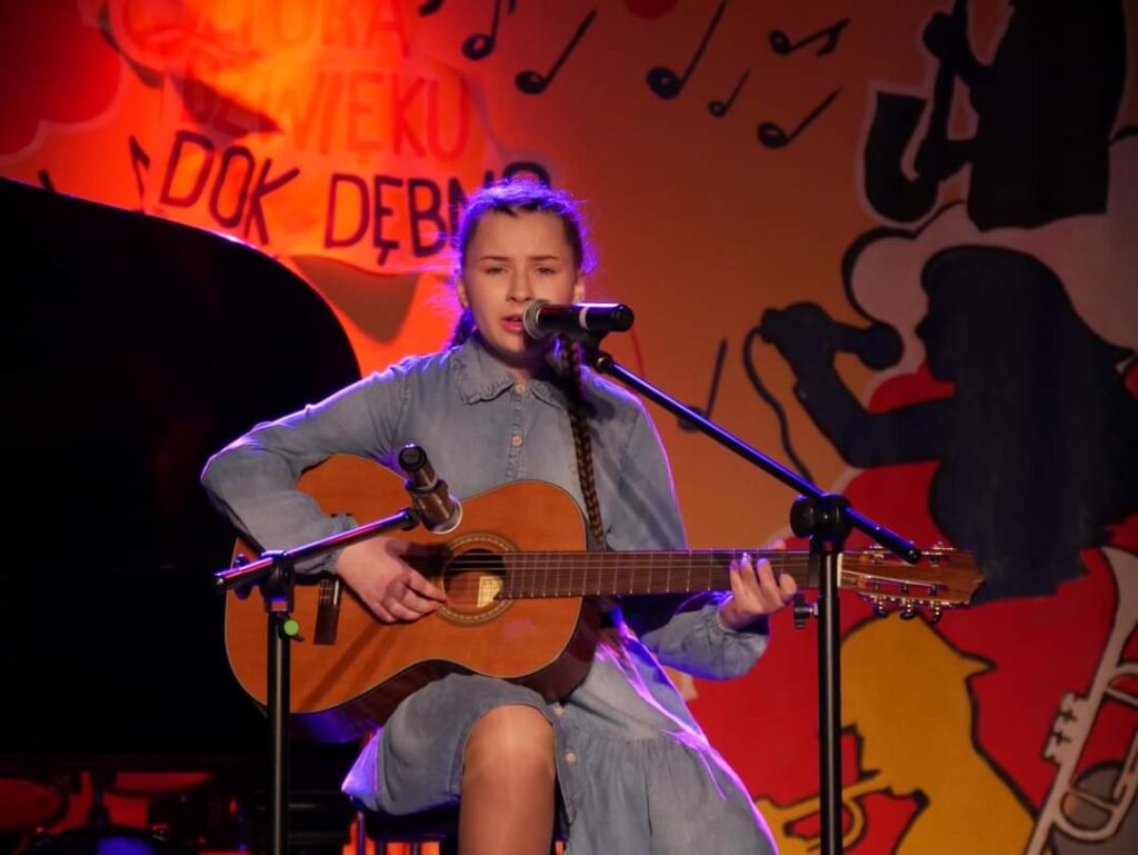 Dziewczynka śpiewa na scenie grając na gitarze klasycznej