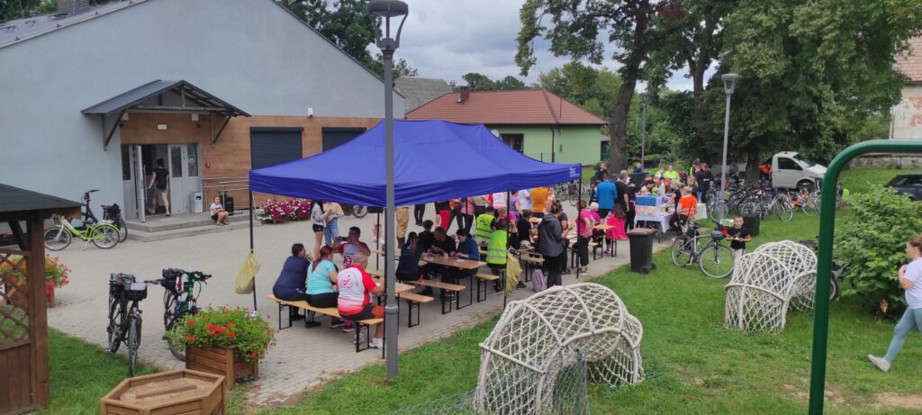 Ławki pod namiotem, świetlica wiejska, teren z kostki brukowej, rowerzyści przy posiłku