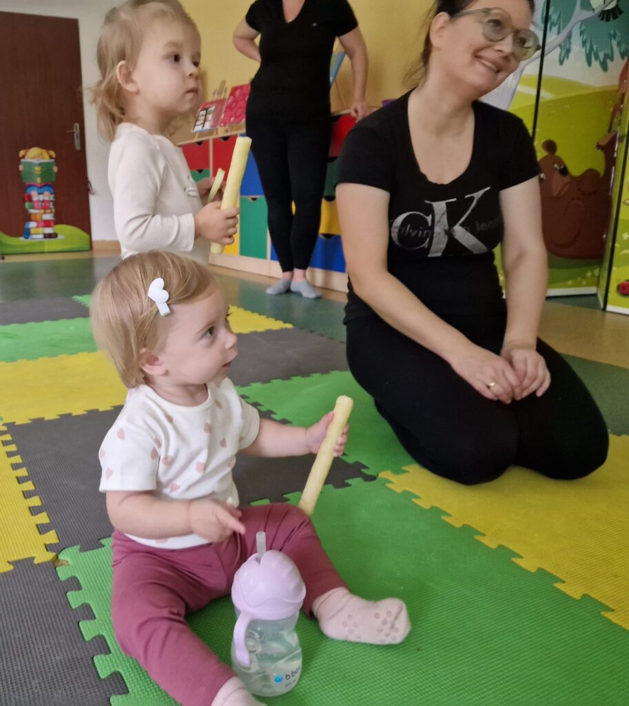Dzieci z opiekunami siedzą na kolorowej macie
