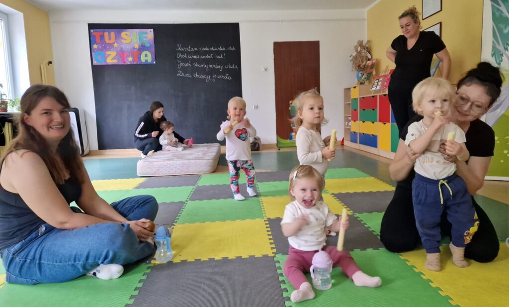 Dzieci z opiekunami siedzą na kolorowej macie
