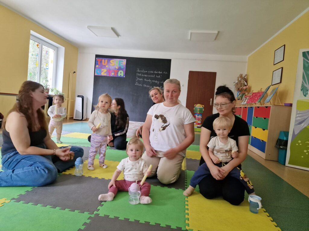 Dzieci z opiekunami siedzą na kolorowej macie