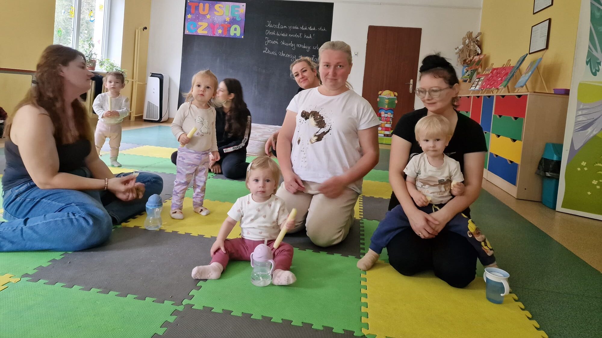 Dzieci z opiekunami siedzą na kolorowej macie