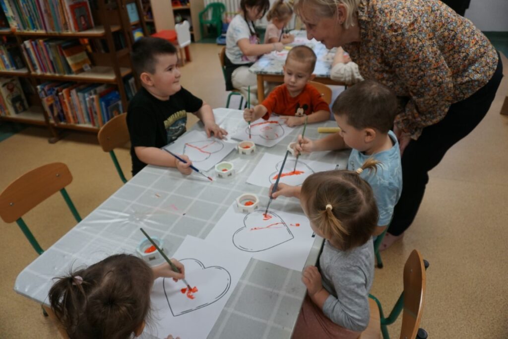 Dzieci z opiekunami, malujące serca farbkami przy stolikach
