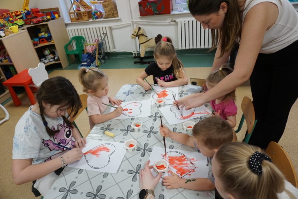 Dzieci z opiekunami, malujące serca farbkami przy stolikach