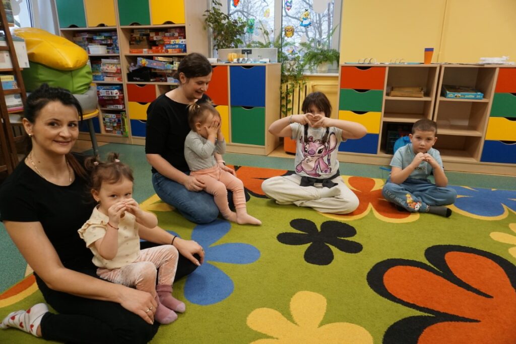 Dzieci z opiekunami siedzące na dywanie, robiące serce z palców