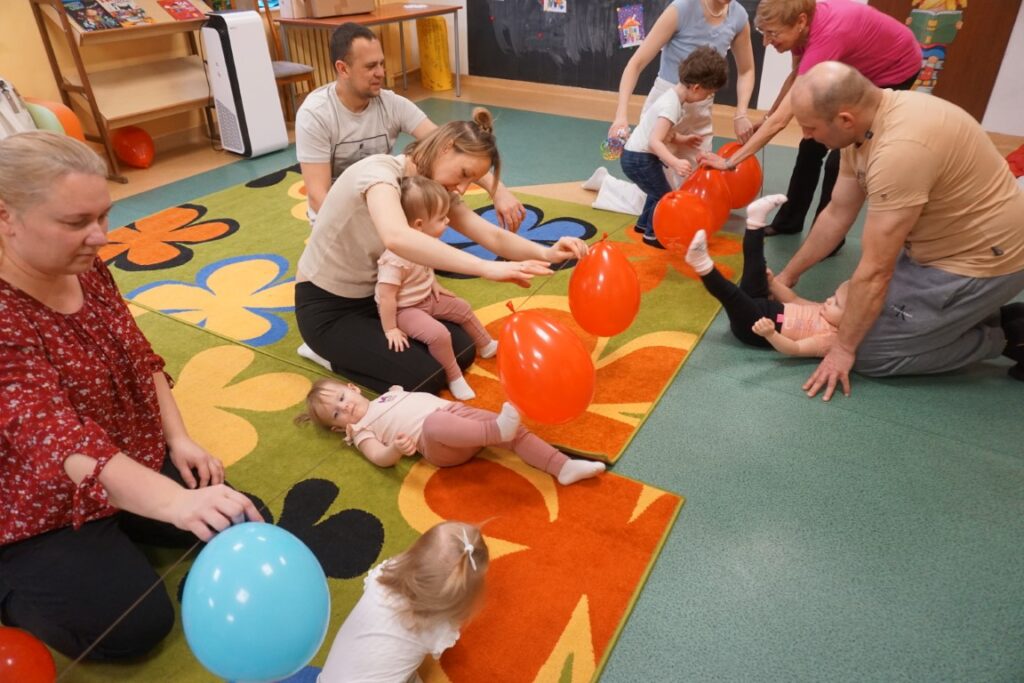 Dzieci siedzące z opiekunami na dywanie i bawiące się kolorowymi balonami
